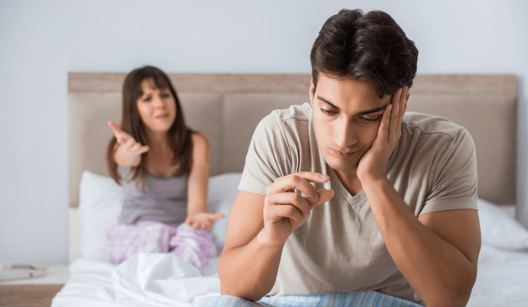 Man holding anti-depressant pill, sitting on bed with partner in the background